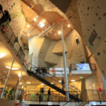 Indoor-Kletterhalle mit hohen Kletterwänden, künstlichen Griffen, Treppen, Geländern und kletternden und gehenden Menschen in einer geräumigen, gut beleuchteten Anlage.