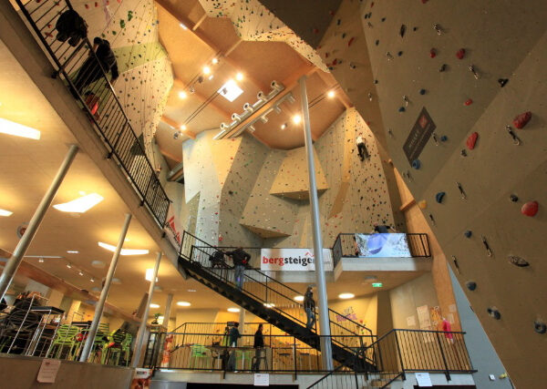 Indoor-Kletterhalle mit hohen Kletterwänden, künstlichen Griffen, Treppen, Geländern und kletternden und gehenden Menschen in einer geräumigen, gut beleuchteten Anlage.