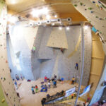 In einer großen, gut beleuchteten Kletterhalle klettern Menschen an verschiedenen Felswänden. Einige Kletterer klettern die Wände hoch, während andere am Boden sitzen und sich vorbereiten oder zuschauen.