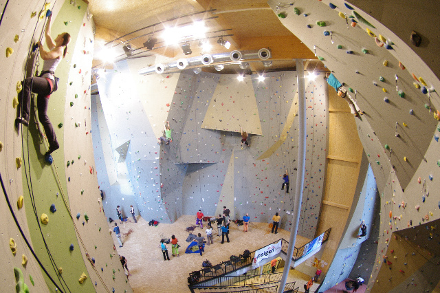 In einer großen, gut beleuchteten Kletterhalle klettern Menschen an verschiedenen Felswänden. Einige Kletterer klettern die Wände hoch, während andere am Boden sitzen und sich vorbereiten oder zuschauen.