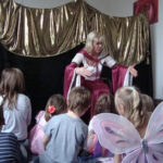 Eine Frau in einem Kostüm erzählt einer Gruppe von kleinen Kindern, die auf dem Boden sitzen, eine Geschichte, wobei ein Kind Schmetterlingsflügel trägt. Hinter ihr befindet sich ein gold-schwarz drapierter Bühnenhintergrund.