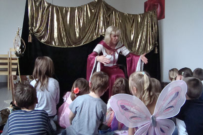 Eine Frau in einem Kostüm erzählt einer Gruppe von kleinen Kindern, die auf dem Boden sitzen, eine Geschichte, wobei ein Kind Schmetterlingsflügel trägt. Hinter ihr befindet sich ein gold-schwarz drapierter Bühnenhintergrund.