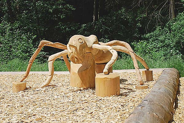 Große Holzskulptur einer Ameise mit sechs Beinen, die auf Holzspänen in einem bewaldeten Außenbereich steht.