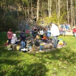 Eine Gruppe von Kindern und Erwachsenen sitzt auf Baumstämmen auf einer von Bäumen umgebenen Wiese und hat sich um ein Lagerfeuer versammelt.