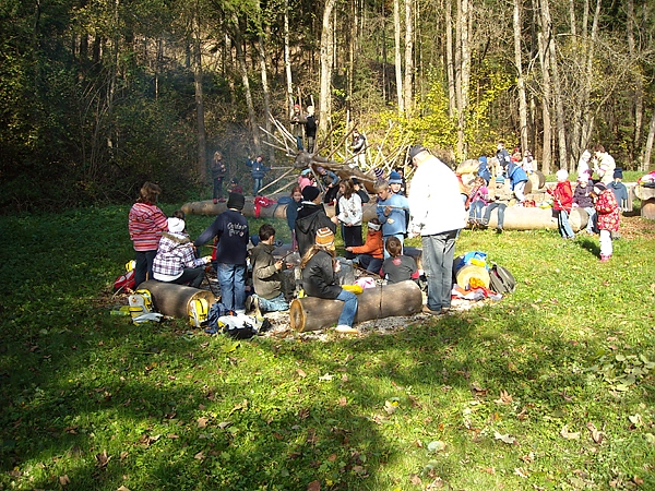 Eine Gruppe von Kindern und Erwachsenen sitzt auf Baumstämmen auf einer von Bäumen umgebenen Wiese und hat sich um ein Lagerfeuer versammelt.