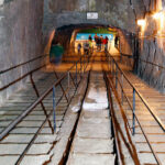 Breiter unterirdischer Tunnel mit Treppen, Handläufen und Schienen in der Mitte; mehrere Personen gehen auf eine beleuchtete Öffnung am anderen Ende zu.