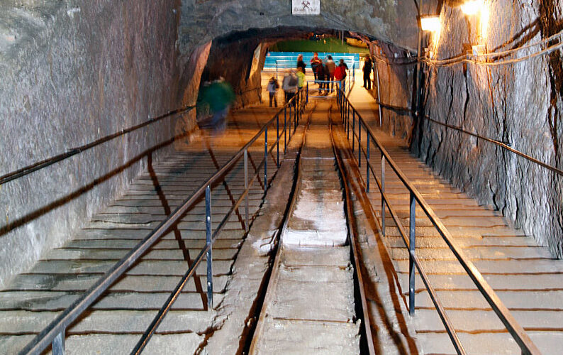 Breiter unterirdischer Tunnel mit Treppen, Handläufen und Schienen in der Mitte; mehrere Personen gehen auf eine beleuchtete Öffnung am anderen Ende zu.