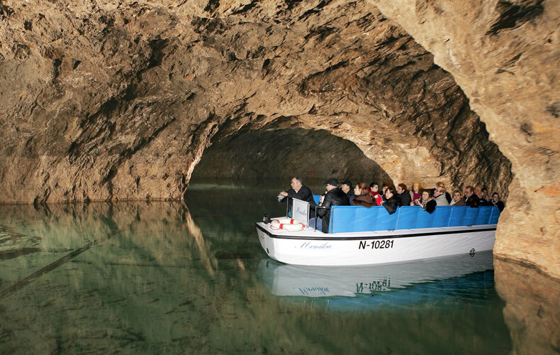 Eine Gruppe von Menschen fährt in einem weißen Boot mit blauen Sitzen durch eine unterirdische Höhle, die mit klarem Wasser gefüllt ist.