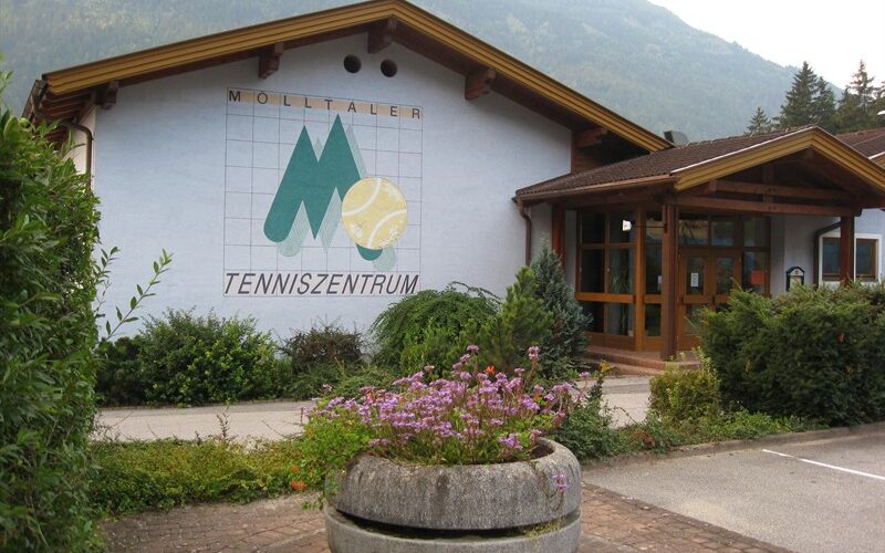 Ein Gebäude mit der Aufschrift "Mölltaler Tenniszentrum" und einem großen Wandgemälde zum Thema Tennis an der Außenwand; im Vordergrund Blumen und Grünpflanzen.
