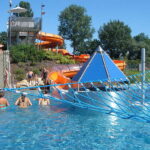 An einem sonnigen Tag schwimmen Menschen in einem Freibad mit einem blauen Kletternetz, einer Pyramidenstruktur und orangefarbenen Wasserrutschen im Hintergrund.