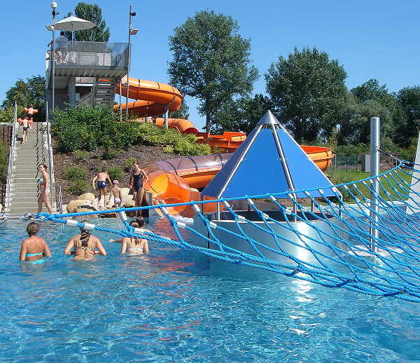 An einem sonnigen Tag schwimmen Menschen in einem Freibad mit einem blauen Kletternetz, einer Pyramidenstruktur und orangefarbenen Wasserrutschen im Hintergrund.