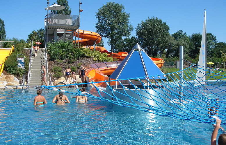An einem sonnigen Tag schwimmen Menschen in einem Freibad mit einem blauen Kletternetz, einer Pyramidenstruktur und orangefarbenen Wasserrutschen im Hintergrund.