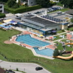 Luftaufnahme eines Freibadkomplexes mit einem Hauptbecken, einer Wasserrutsche, Liegeflächen und einem angrenzenden Gebäude, umgeben von Rasen und einem Parkplatz.