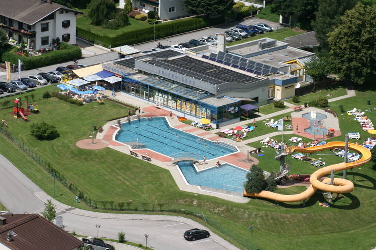 Luftaufnahme eines Freibadkomplexes mit einem Hauptbecken, einer Wasserrutsche, Liegeflächen und einem angrenzenden Gebäude, umgeben von Rasen und einem Parkplatz.