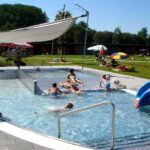 Kinder und Erwachsene schwimmen und spielen in der Nähe einer blauen Rutsche in einem Außenpool mit Sonnenschirmen und Rasenflächen.