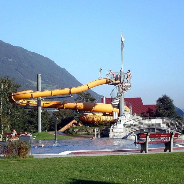 Wasserpark im Freien mit einer gelben Spiralwasserrutsche, einem kleinen Becken, Spaziergängern und Bergen im Hintergrund an einem klaren Tag.