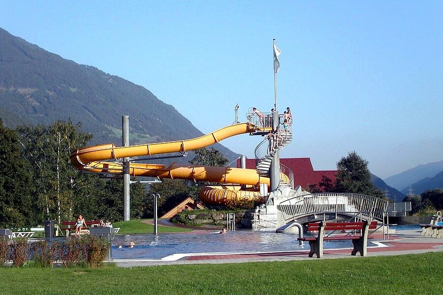 Wasserpark im Freien mit einer gelben Spiralwasserrutsche, einem kleinen Becken, Spaziergängern und Bergen im Hintergrund an einem klaren Tag.