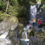 Vier Personen stehen und knien auf Felsen neben einem Wasserfall, umgeben von üppig grünen Bäumen und moosbewachsenen Steinen.