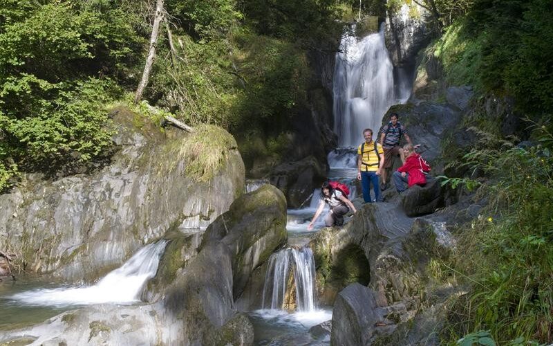 Vier Personen stehen und knien auf Felsen neben einem Wasserfall, umgeben von üppig grünen Bäumen und moosbewachsenen Steinen.