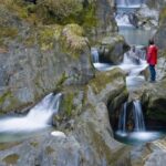 Eine Person in einer roten Jacke steht auf einem Felsen neben einem klaren, fließenden Bach mit kleinen Wasserfällen in einem felsigen, bewaldeten Gebiet.