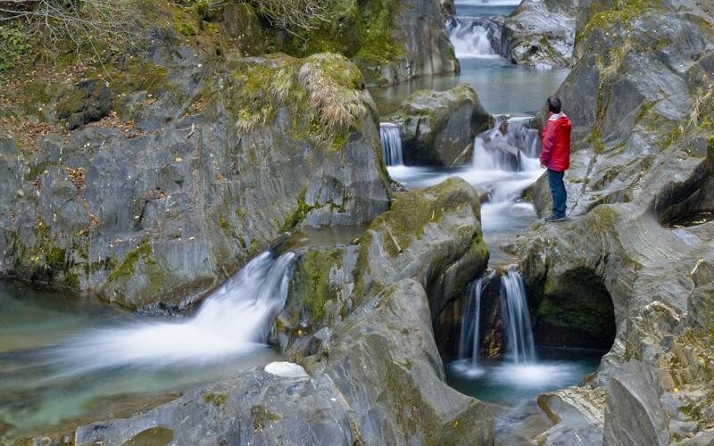 Eine Person in einer roten Jacke steht auf einem Felsen neben einem klaren, fließenden Bach mit kleinen Wasserfällen in einem felsigen, bewaldeten Gebiet.