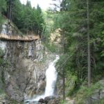 Ein Holzsteg führt entlang einer Klippe oberhalb eines felsigen Wasserfalls, umgeben von dichten grünen Kiefern in einem Waldgebiet.