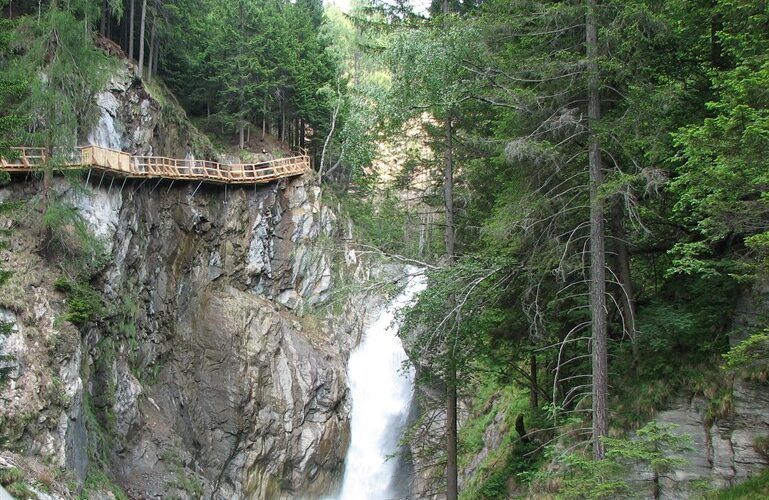 Ein Holzsteg führt entlang einer Klippe oberhalb eines felsigen Wasserfalls, umgeben von dichten grünen Kiefern in einem Waldgebiet.