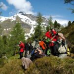 Eine Gruppe von Menschen auf einer grasbewachsenen Anhöhe mit einem Fernglas, im Hintergrund Kiefern und schneebedeckte Berge.