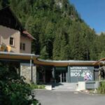 Ein Gebäude mit der Aufschrift "Nationalparkzentrum BIOS" steht an einem bewaldeten Berghang unter einem klaren Himmel.