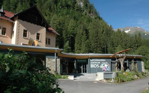 Ein Gebäude mit der Aufschrift "Nationalparkzentrum BIOS" steht an einem bewaldeten Berghang unter einem klaren Himmel.