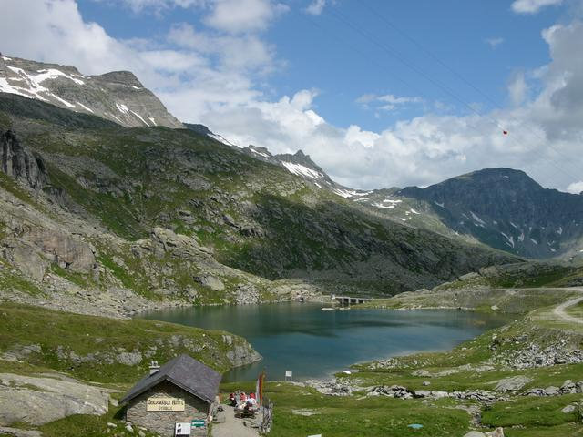 Eine Berglandschaft mit einem kleinen See, felsigen grünen Hügeln, ein paar Schneeflecken, einer Hütte im Vordergrund und einem teilweise bewölkten Himmel.