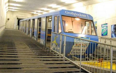 Ein blauer Standseilbahnwagen mit der Aufschrift "Großglockner" steht an einer steilen Steigung in einem Betontunnel, der auf beiden Seiten mit Geländern versehen ist.