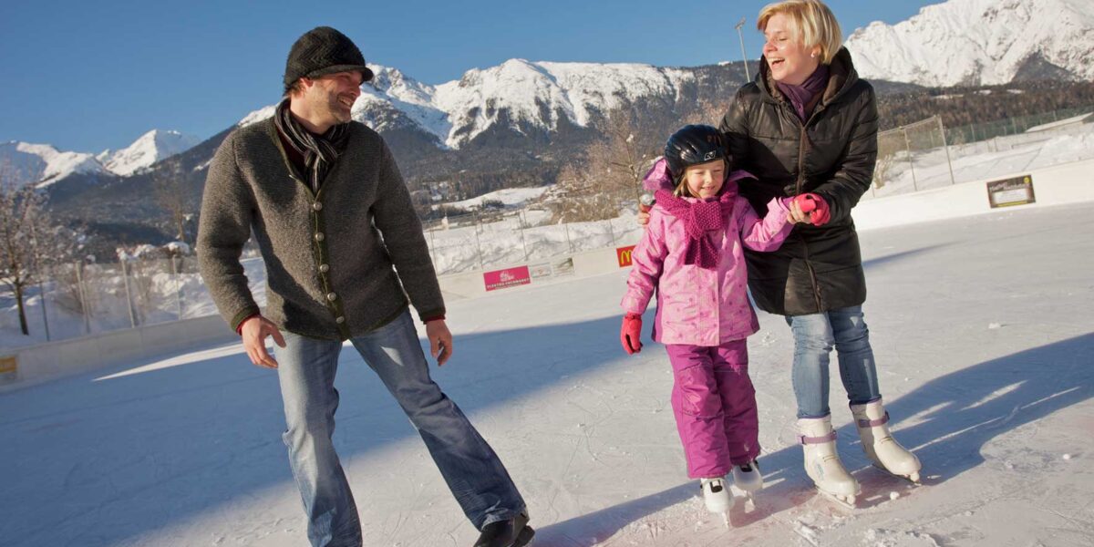 Drei Personen, zwei Erwachsene und ein Kind, beim Schlittschuhlaufen im Freien an einem sonnigen Tag mit schneebedeckten Bergen im Hintergrund.