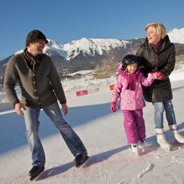 Drei Personen, zwei Erwachsene und ein Kind, beim Schlittschuhlaufen im Freien an einem sonnigen Tag mit schneebedeckten Bergen im Hintergrund.