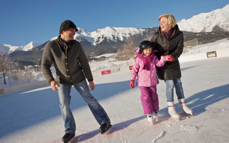 Drei Personen, zwei Erwachsene und ein Kind, beim Schlittschuhlaufen im Freien an einem sonnigen Tag mit schneebedeckten Bergen im Hintergrund.