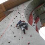 Eine Person klettert an einer künstlichen Felswand im Freien mit Hilfe von Griffen und Tritten, wobei ein Sicherungsseil über dem Kopf angebracht ist.