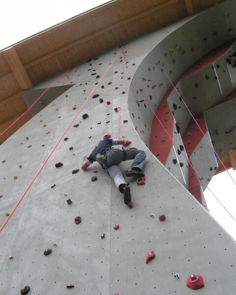 Eine Person klettert an einer künstlichen Felswand im Freien mit Hilfe von Griffen und Tritten, wobei ein Sicherungsseil über dem Kopf angebracht ist.