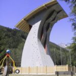 Eine hohe Kletterwand mit überhängendem Dach, umgeben von einem Holzzaun, steht in der Nähe eines Spielplatzes und von Bäumen unter freiem Himmel.