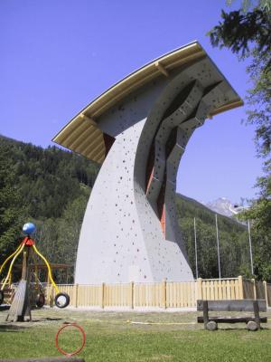 Eine hohe Kletterwand mit überhängendem Dach, umgeben von einem Holzzaun, steht in der Nähe eines Spielplatzes und von Bäumen unter freiem Himmel.