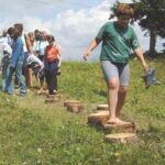 Eine Gruppe von Kindern und Erwachsenen läuft barfuß auf hölzernen Trittsteinen, die in einem grasbewachsenen Außenbereich unter einem teilweise bewölkten Himmel angeordnet sind.