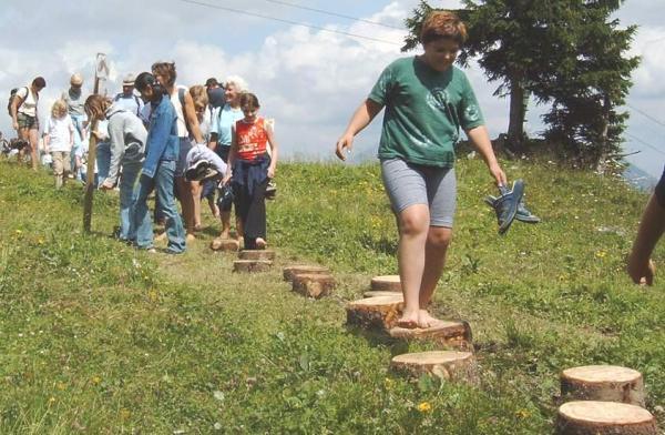 Eine Gruppe von Kindern und Erwachsenen läuft barfuß auf hölzernen Trittsteinen, die in einem grasbewachsenen Außenbereich unter einem teilweise bewölkten Himmel angeordnet sind.