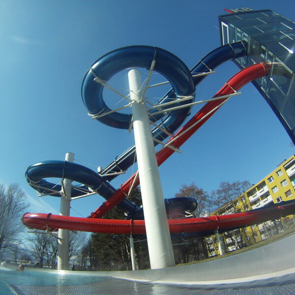 Wasserrutschenkomplex im Freien mit blauen und roten Rutschen, die sich um weiße Stützen winden, neben einem Glasgebäude und Bäumen unter einem klaren blauen Himmel.
