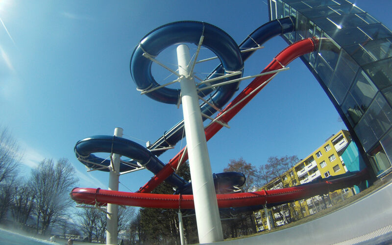 Wasserrutschenkomplex im Freien mit blauen und roten Rutschen, die sich um weiße Stützen winden, neben einem Glasgebäude und Bäumen unter einem klaren blauen Himmel.