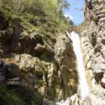 Ein hoher Wasserfall fließt eine von Bäumen umgebene Felswand hinunter, während links zwei Wanderer auf einem Pfad stehen.