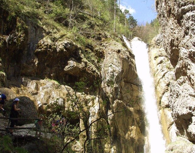 Ein hoher Wasserfall fließt eine von Bäumen umgebene Felswand hinunter, während links zwei Wanderer auf einem Pfad stehen.