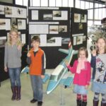 Vier Kinder stehen in einem Museum vor einem Modellflugzeug und Schautafeln, jedes hält ein kleines Flugzeug mit Gummibandantrieb in der Hand.