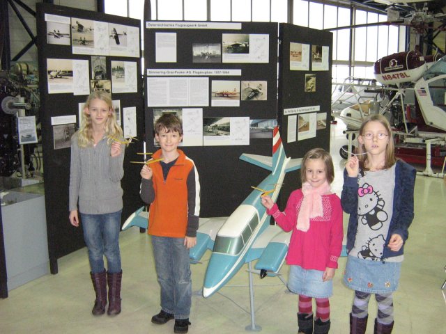 Vier Kinder stehen in einem Museum vor einem Modellflugzeug und Schautafeln, jedes hält ein kleines Flugzeug mit Gummibandantrieb in der Hand.