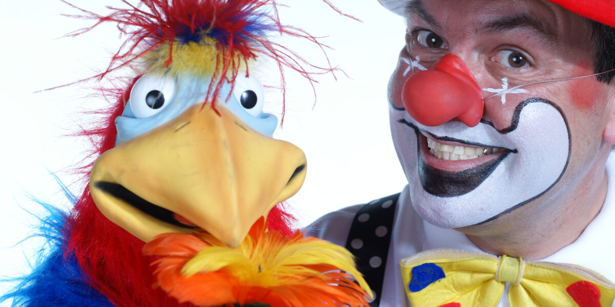 Ein als Clown verkleideter Mann mit geschminktem Gesicht und roter Nase steht neben einer bunten Vogelpuppe, die eine gelbe Blume hält.