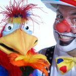 Ein als Clown verkleideter Mann mit geschminktem Gesicht und roter Nase steht neben einer bunten Vogelpuppe, die eine gelbe Blume hält.