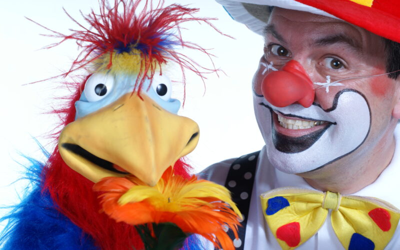 Ein als Clown verkleideter Mann mit geschminktem Gesicht und roter Nase steht neben einer bunten Vogelpuppe, die eine gelbe Blume hält.